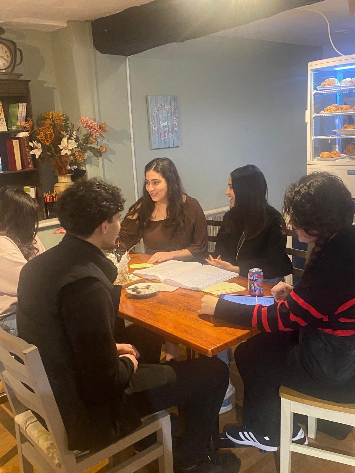 A Bible study at Hesed: five people gathered round a wooden table with an open Bible and notes in front of them, talking and laughing together, with bookshelves, flowers and the cake fridge behind.