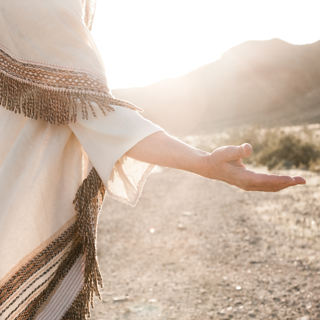 A figure in a cream robe standing in bright sunlight on a desert path, one hand held open in welcome.