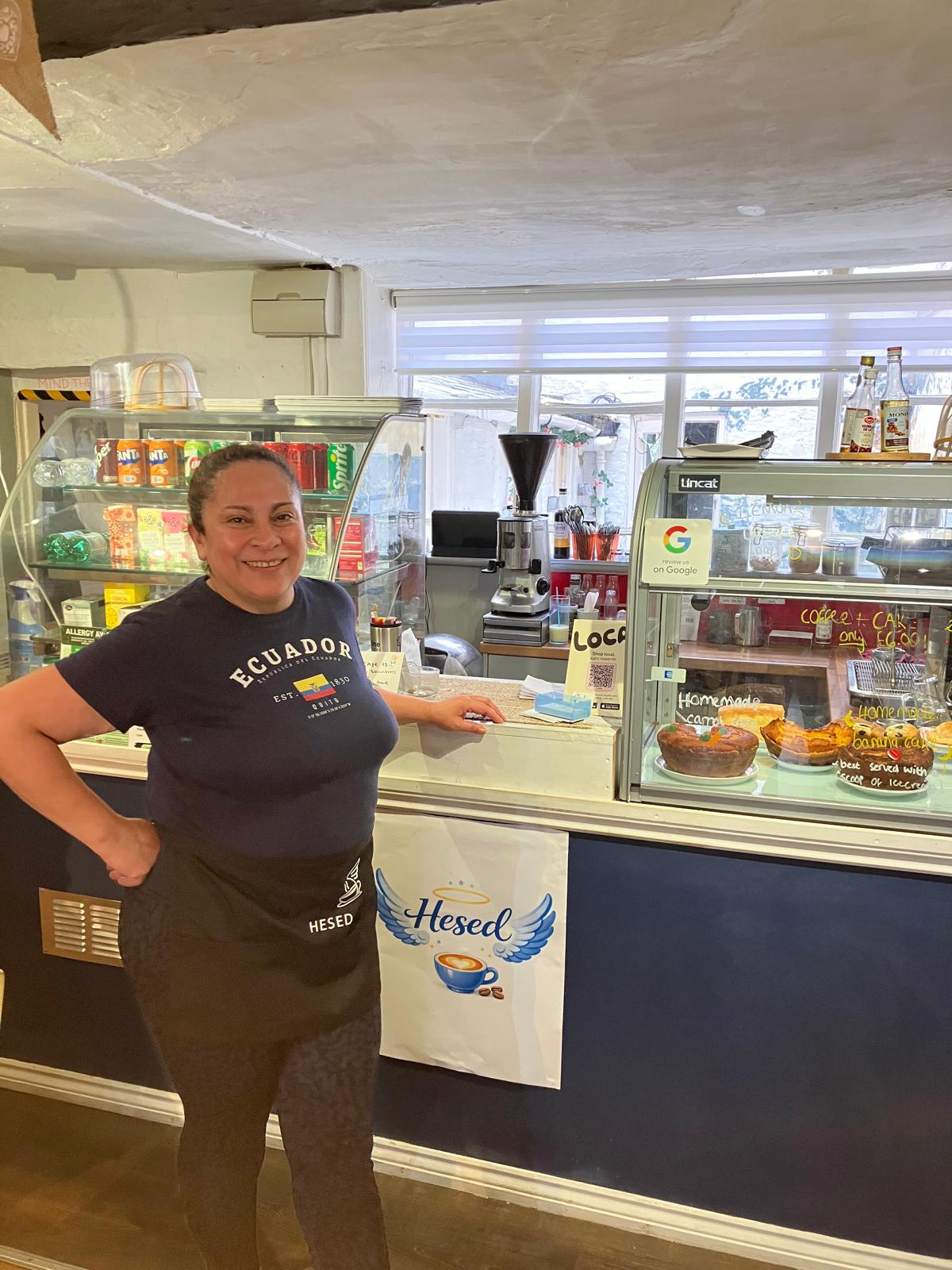 Jhoced standing beside the counter at Hesed, smiling, in an Ecuador T-shirt and a Hesed apron, with the winged-cup Hesed sign on the counter front and a cabinet of homemade cakes behind her.