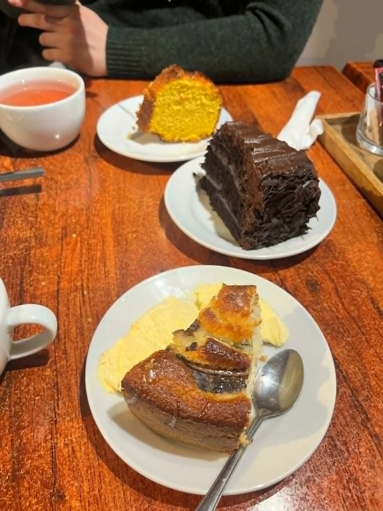 Three cakes on white plates on a wooden table — a slice of orange sponge, a slice of dark chocolate layer cake, and banana bread with custard — beside a cup of tea.