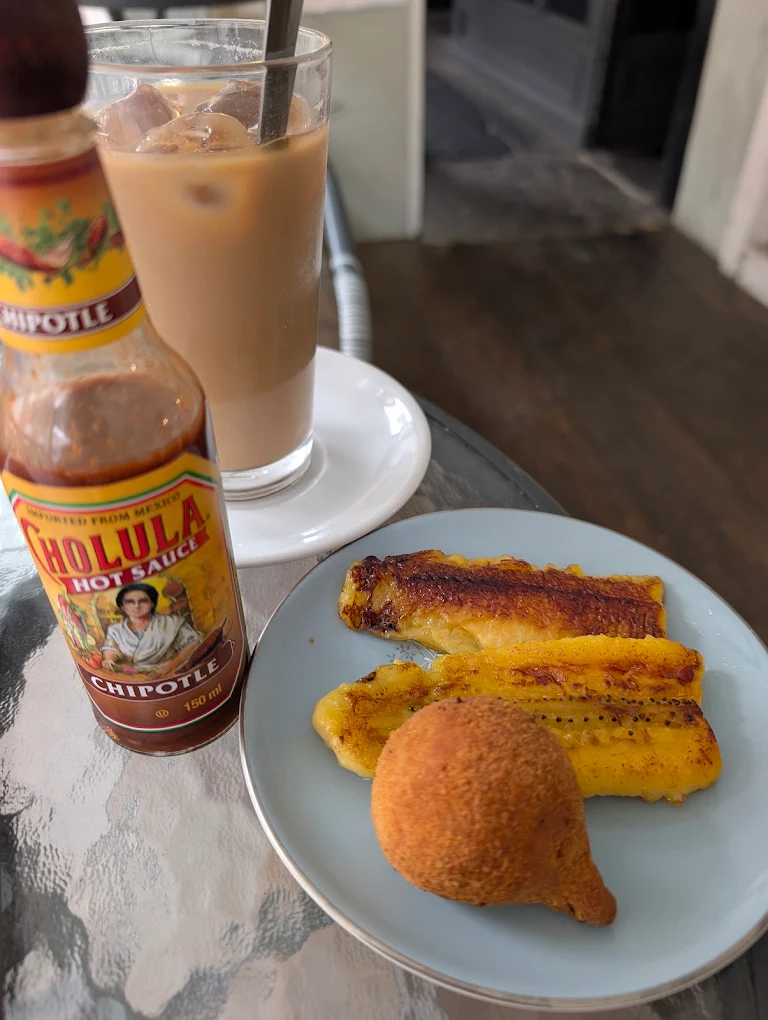 A pale blue plate of griddled sweet plantains and a golden coxinha, with an iced coffee and a bottle of chipotle hot sauce alongside.