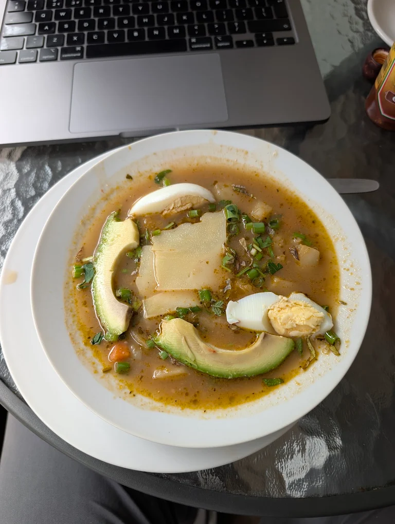 A white bowl of Ecuadorian soup with sliced potato, avocado, boiled egg and spring onions.