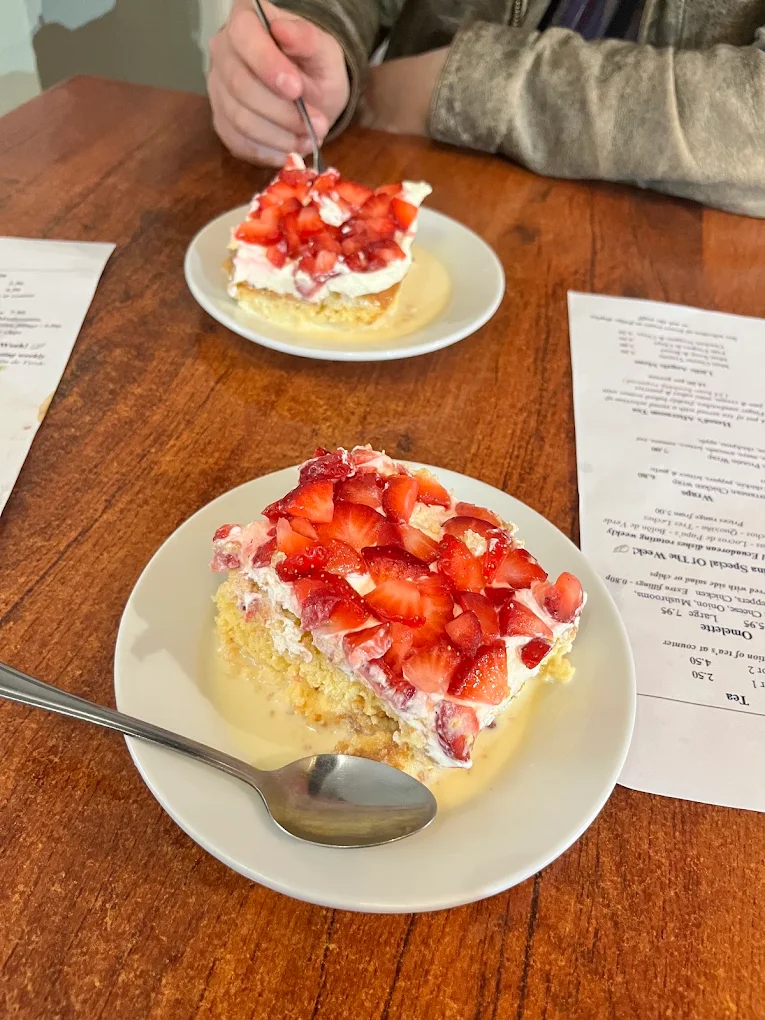 A slice of tres leches cake soaked in milk, topped with cream and chopped strawberries, on a white plate.