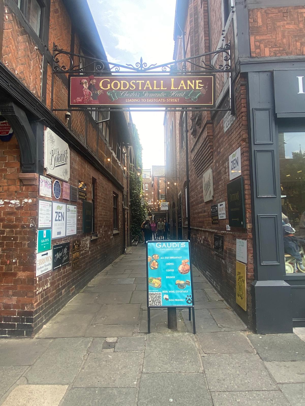The entrance to Godstall Lane in Chester: a narrow paved lane between red brick buildings, with an ornate iron sign overhead reading &ldquo;Godstall Lane — Chester&rsquo;s Romantic Heart, leading to Eastgate Street.&rdquo;
