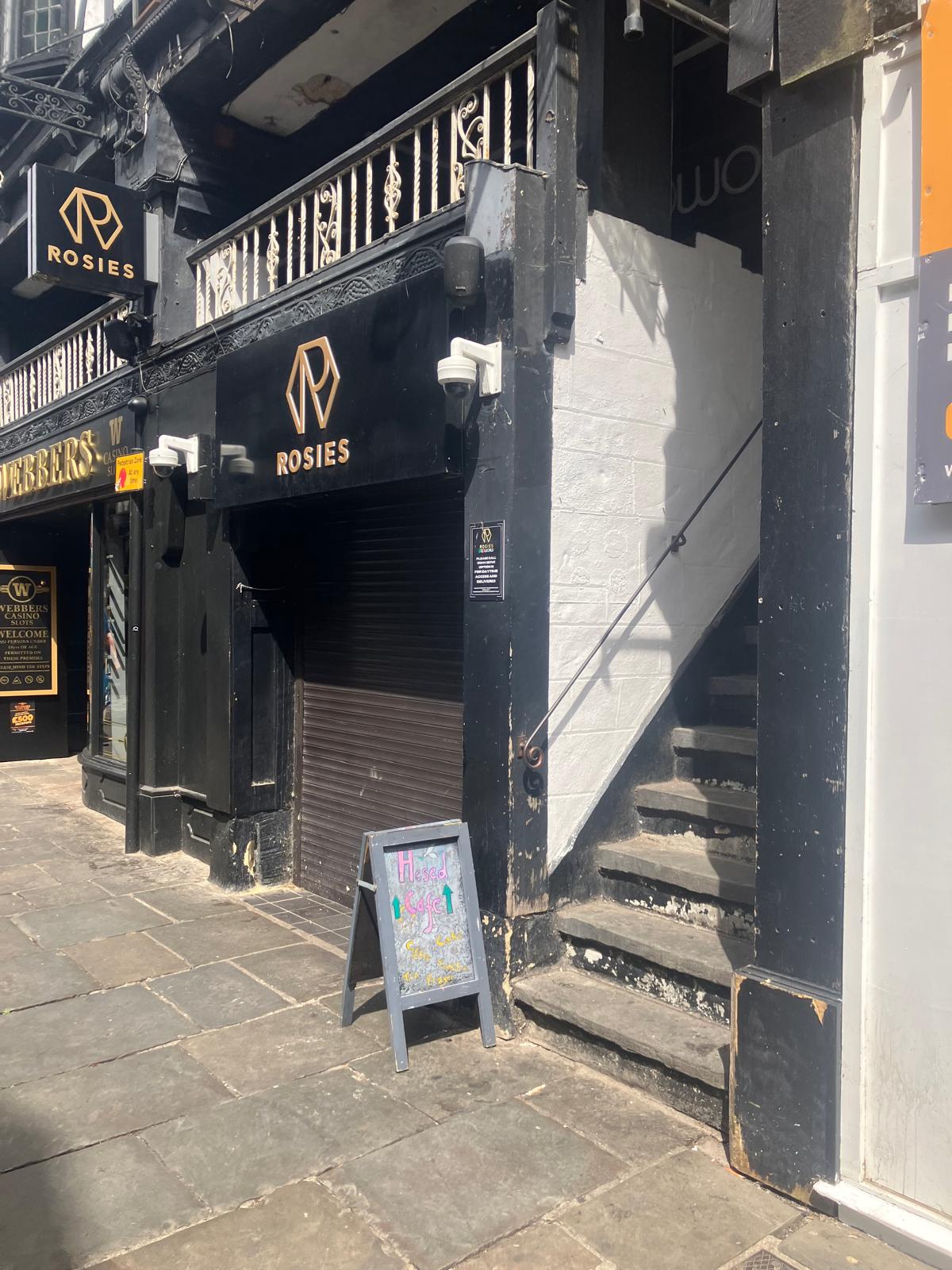The street entrance in Chester: a black timber-framed frontage with a chalkboard A-board reading &ldquo;Hesed Café — Coffee, Cake, Prayer&rdquo; at the foot of a flight of stone steps leading up to the Rows.