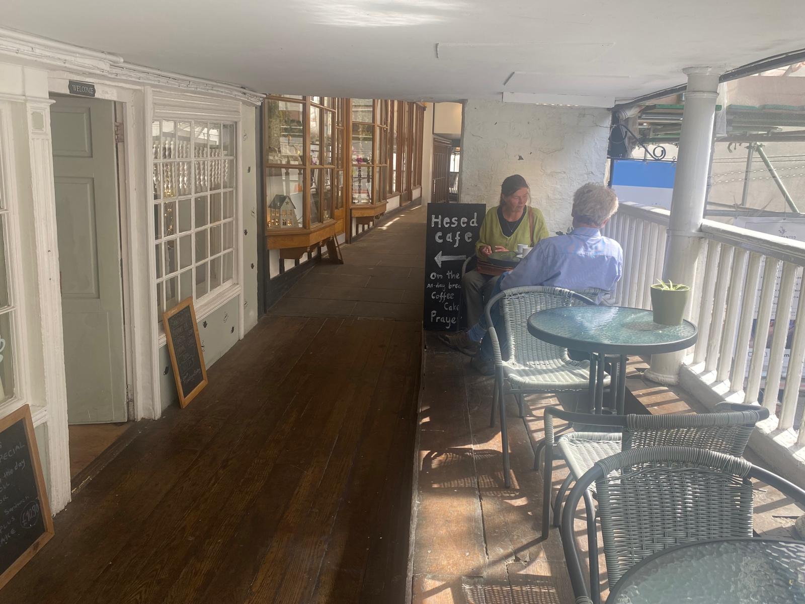 The covered walkway of the Chester Rows: a wooden gallery with shopfronts along one side, tables and chairs along the other, and a chalkboard sign pointing the way to Hesed Café for all-day breakfast, coffee, cake and prayer.